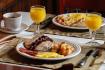 Table with plates with pancakes, eggs, potatoes and bacon along with a glass of orange juice and a cup of coffee